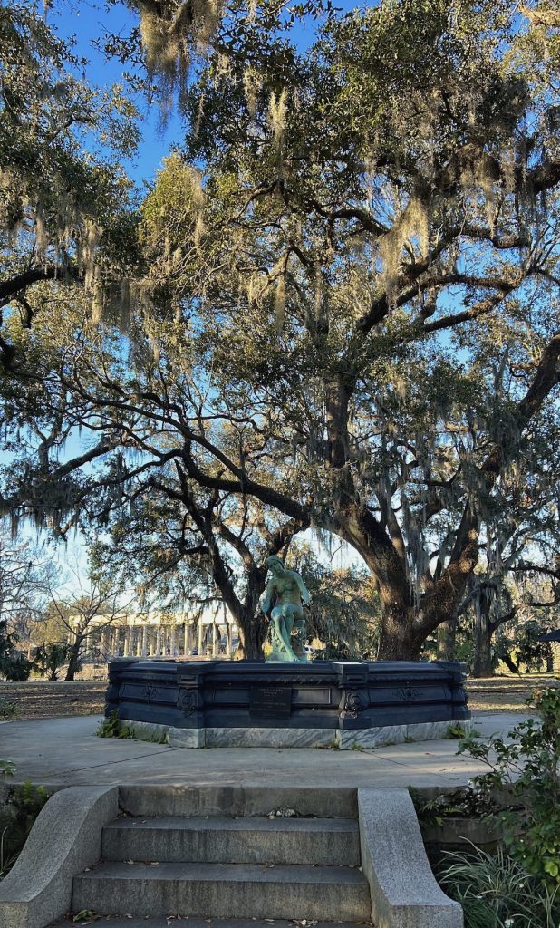 A statue of a woman in a turned off fountain with trees, branches and blue skies behind her in New Orleans' City Park