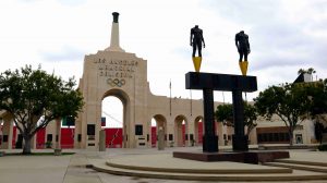The peristyle of the Los Angeles Memorial Coliseum in the background and a statue of a male and female olympian in the foreground.