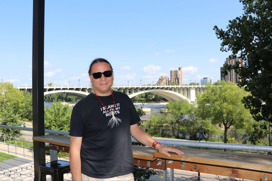 Man wearing sunglasses stands on patio with bridge and city skyline backdrop