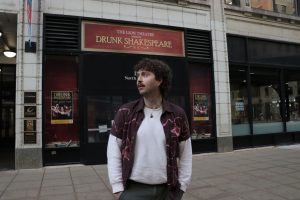 Caleb Harbin stands in front of a “Drunk Shakespeare” sign that hangs at The Lion Theatre.