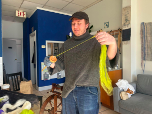 Man in a gray sweater working with a spinning wheel and yellow yarn