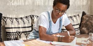 Fashionable dark skinned girl studies chart, works with many documents, writes down in notebook
