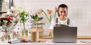 Young flower shop owner using laptop at counter. Woman florist standing at table.