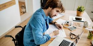 Man writing on paper Young business owner in wheelchair working on papers at table with laptop