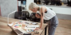 Young stylish blonde tattooed female designer making some sketches and working with color swatch samples while standing near office desk