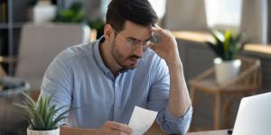 Male small business owner with stressed expression while looking at laptop and holding receipt