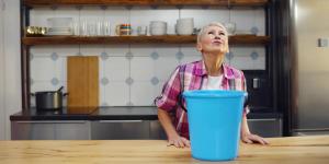 Worried aged woman holding bucket while water droplets leak from ceiling in kitchen