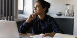 Woman thinking in office Woman sitting at table thinking with laptop in front of her in home