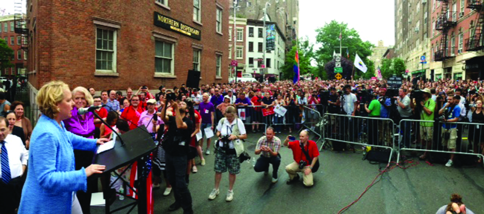 Image shows two people at a podium in front of a large crowd on a city street. One person speaks to the crowd, while the other stands next to the podium.