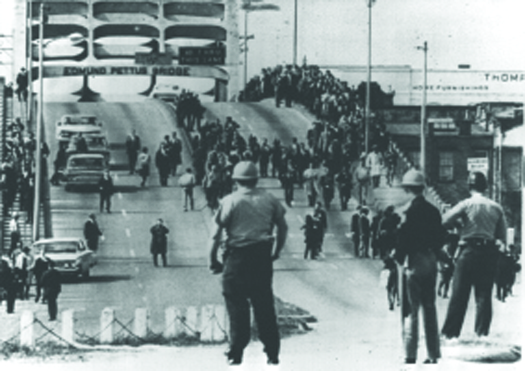 An image of a bridge. On the right of the overpass are several people marching in a large crowd. In the foreground are uniformed people watching the marchers.