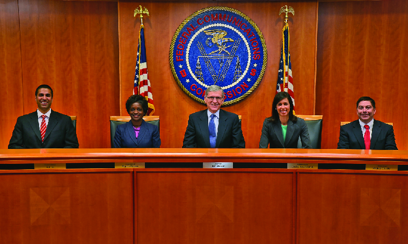 An image from left to right of Ajit Pai, Mignon Clyburn, Chairman Tom Wheeler, Jessica Rosenworcel and Michael O’Rielly seated in front of a large circular banner reading “Federal Communications Commission”.