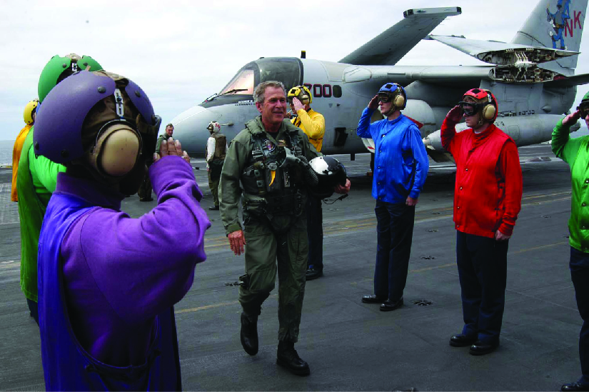 A photo of George W. Bush in a flight suit stepping out of a plane onto an aircraft carrier. Personnel stand on either side and salute him.