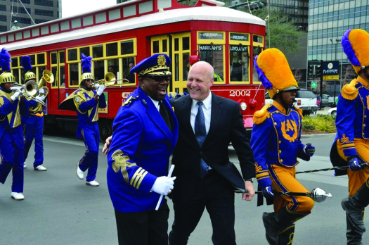 An image of Mitch Landrieu standing in the middle of a group of people who are playing various instruments. A streetcar is in the background.