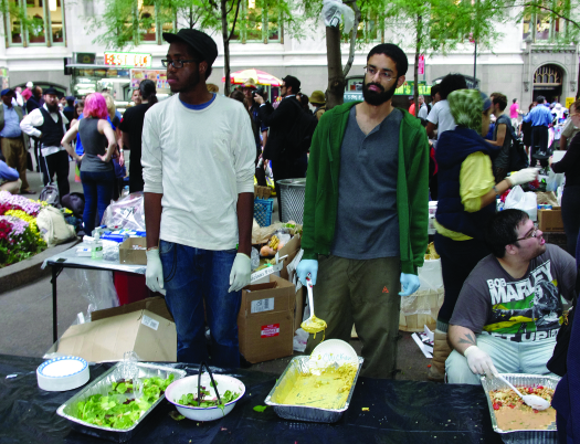 An image of three people behind a table. On the table are serval large open containers of food. A crowd of people is in the background.