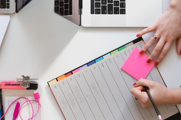 Planejamento de redes sociais: imagem de uma mesa com notebook aberto, uma agenda e uma mão de mulher escrevendo no um post it cor de rosa.
