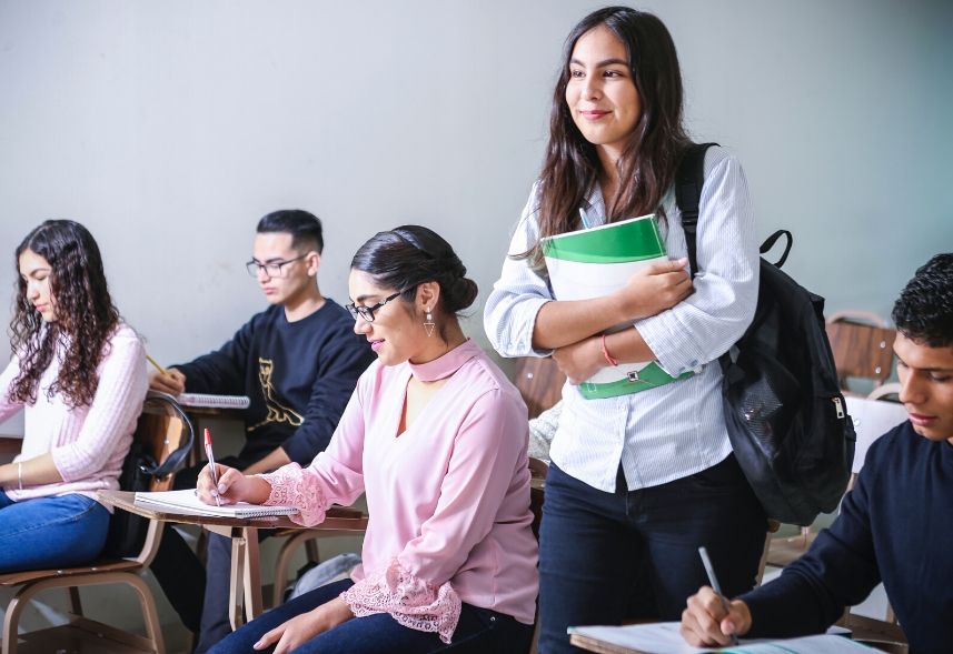 Como fidelizar alunos na escola: imagem de uma sala de aula com vários alunos sentados estudando e uma aluna em pé segurando o material