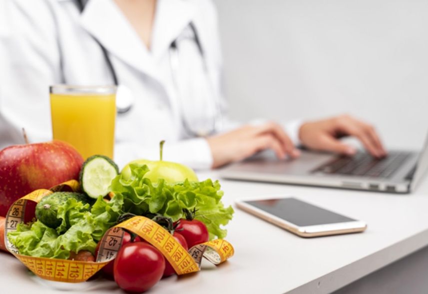 marketing para nutricionista: imagem de um mulher digitando no notebook ao fundo e uma seleção de frutas e verduras e um suco de laranja com uma fita métrica na frente