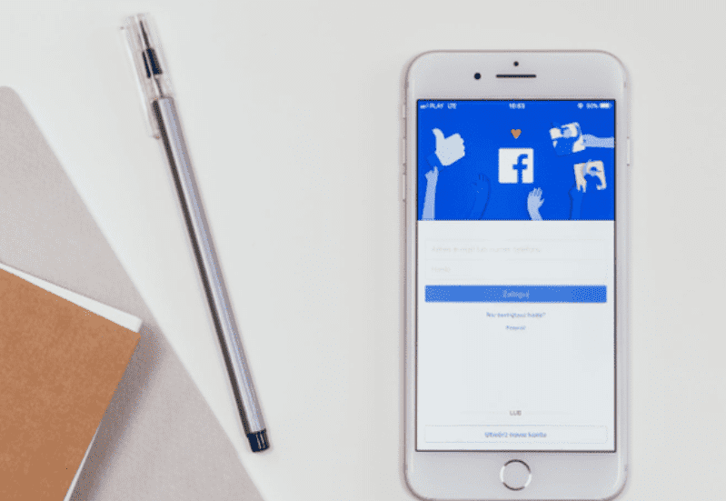 Facebook Analytics header: um celular branco está jogado sobre a mesa, em sua tela imagem do acesso ao Facebook. Ao lado esquerdo, uma caneta também está jogada na diagonal.
