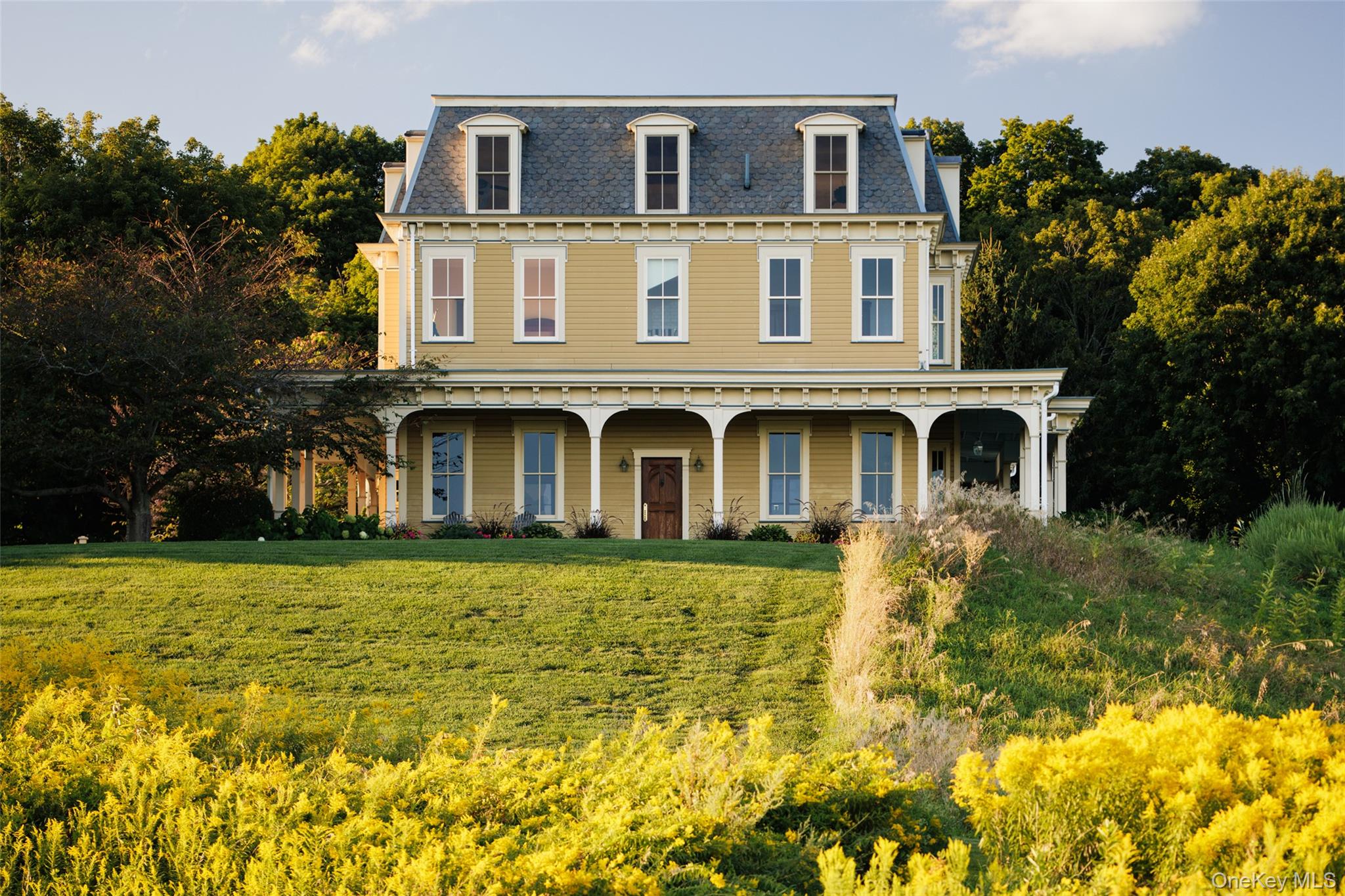 Just a mile and a half south of Millbrook Village, along the scenic stretch of Route 82 where farmland unfolds and county views stretch for miles, a tree-lined driveway winds back from the road to reveal a distinguished country estate. Built in the mid-1990s yet styled with timeless character, this Second Empire Victorian home showcases a sweeping wraparound porch, a classic mansard roof, and intricate dentil molding that capture a bygone elegance. Robust double entry doors open to a grand center hall with soaring 12-foot ceilings. To the right, an office frames sweeping views of the surrounding farmland; to the left, a gracious living room centers on a wood-burning fireplace. A cozy den offers a retreat for quiet evenings, while the formal dining room flows seamlessly to the back patioâ€”perfect for al fresco gatherings. The spacious kitchen features abundant cabinetry, an island, and sun-filled windows framing uninterrupted green vistas. Upstairs, the primary suite includes an ensuite bath, complemented by two additional bedrooms, a full bath, and a second office. A third-floor bonus room offers endless versatilityâ€”ideal for a media lounge, playroom, gym, or game spaceâ€”all surrounded by panoramic views. Set on over five acres, the property is complete with a three-car garage, fenced garden, and meandering walking paths. Less than 90 miles from Manhattan, this home offers the best of country living with easy access to Millbrookâ€™s beloved amenities, including retail and antique shopping, restaurants like Cafe Les Baux, and the much-talked-about House of Stefas, and activities like Innisfree Garden and the Cary Institute.