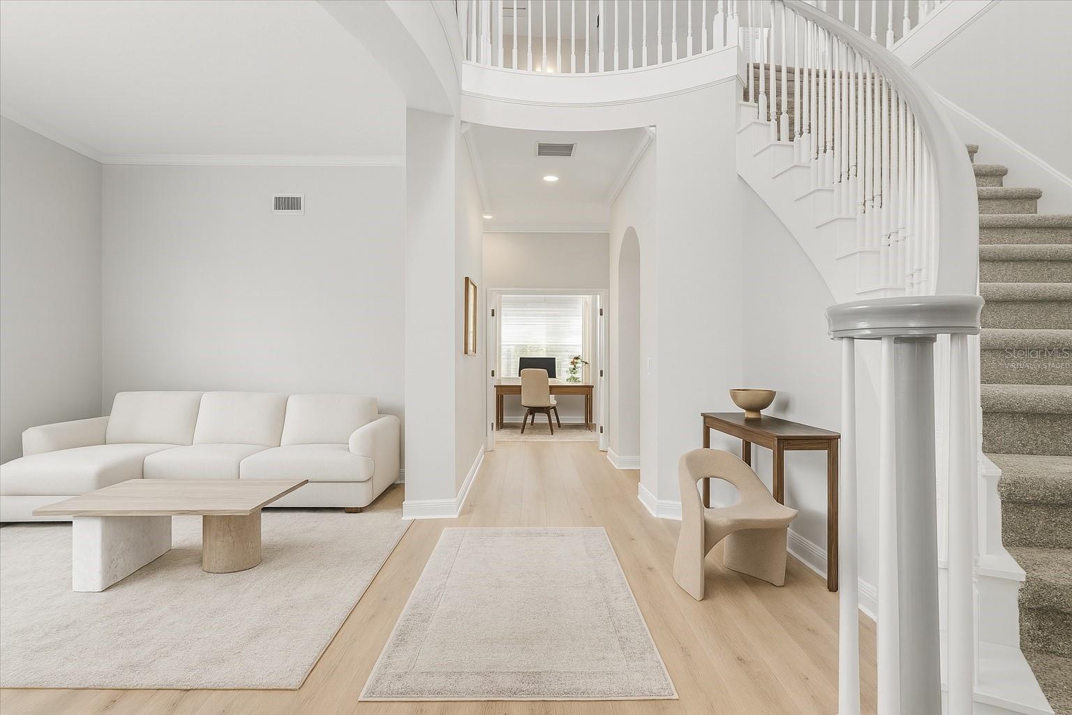 Main entrance hallway with lounge sitting area and rolling center staircase ; hallway to in-law quarters / office in rear of home and virtually staged