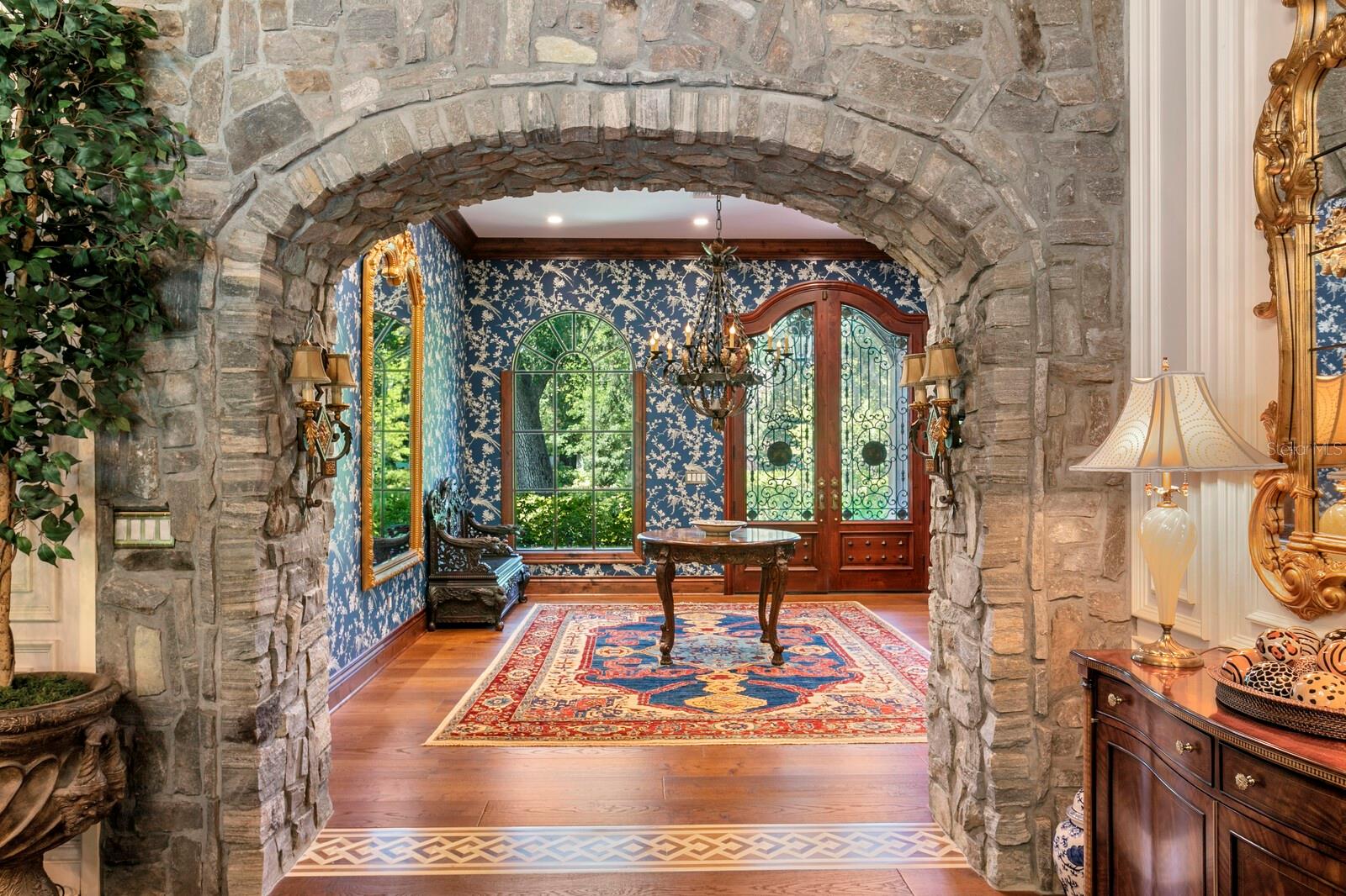 This grand entrance, framed by a rugged stone archway, offers a whimsical peek into a sophisticated haven adorned with intricate wallpaper and timeless wooden accents.