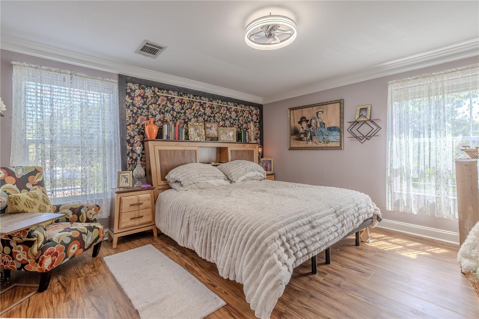 The second bedroom offers comfort, charm, and abundant natural light, making it an ideal guest suite or peaceful personal retreat.Warm wood-look vinyl flooring, crown molding, and large windows create an airy, inviting feel. A whimsical floral accent wall adds a pop of personality behind the bed, while ample space allows for both bedroom furniture and a cozy reading nook. Bright, stylish, and serene—this room is designed for rest and relaxation.