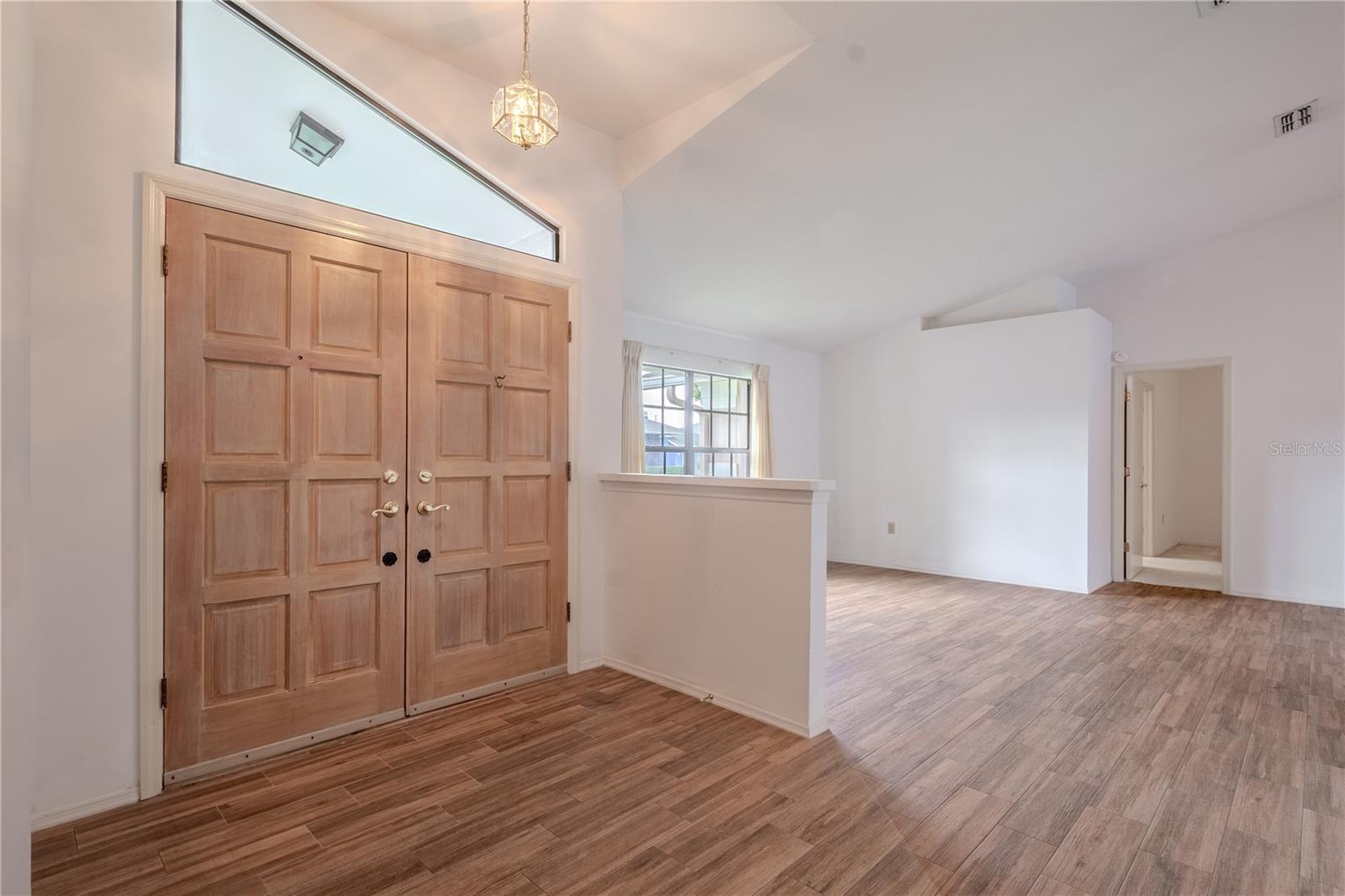 Stately double doors open into a bright, welcoming foyer where classic design meets modern comfort. The rich, wood-look tile flooring offers durability with the warmth of natural grain,  that flows throughout the home. Above, a geometric glass pendant light adds a touch of charm while the transom window overhead invites soft natural light into the space.