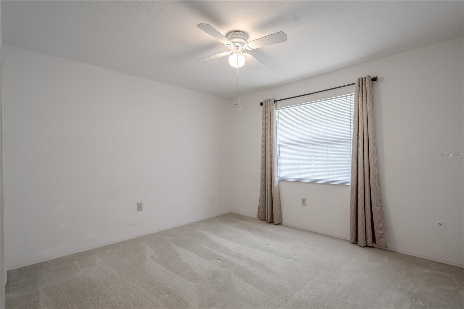 Bedroom 3 is a comfortable and adaptable space with a quiet ambiance and plenty of natural light. A wide window and ceiling fan create a breezy, relaxed feel, while the neutral walls and flooring offer a blank canvas for any design style