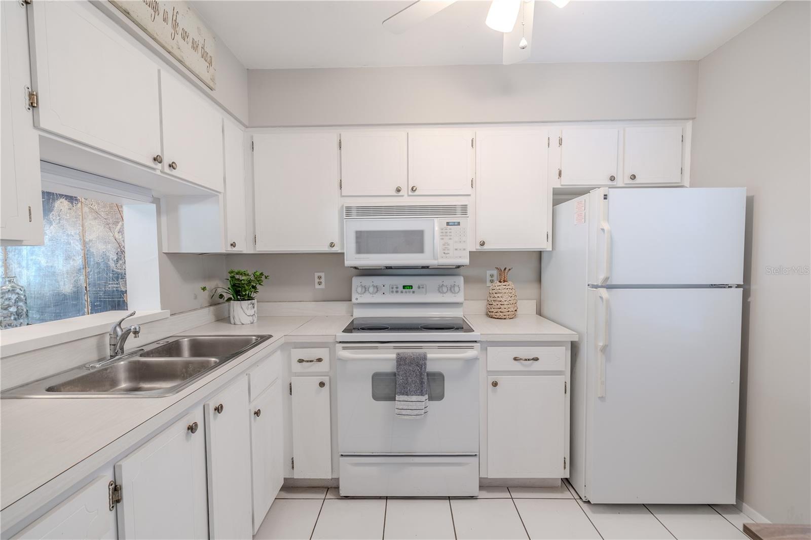 Bright, clean, and efficiently designed, this  kitchen offers timeless appeal with crisp cabinetry, matching white appliances, and sleek neutral tile flooring. The layout maximizes counter space, featuring a double stainless steel sink, smooth-top range, built-in microwave, and full-size refrigerator.