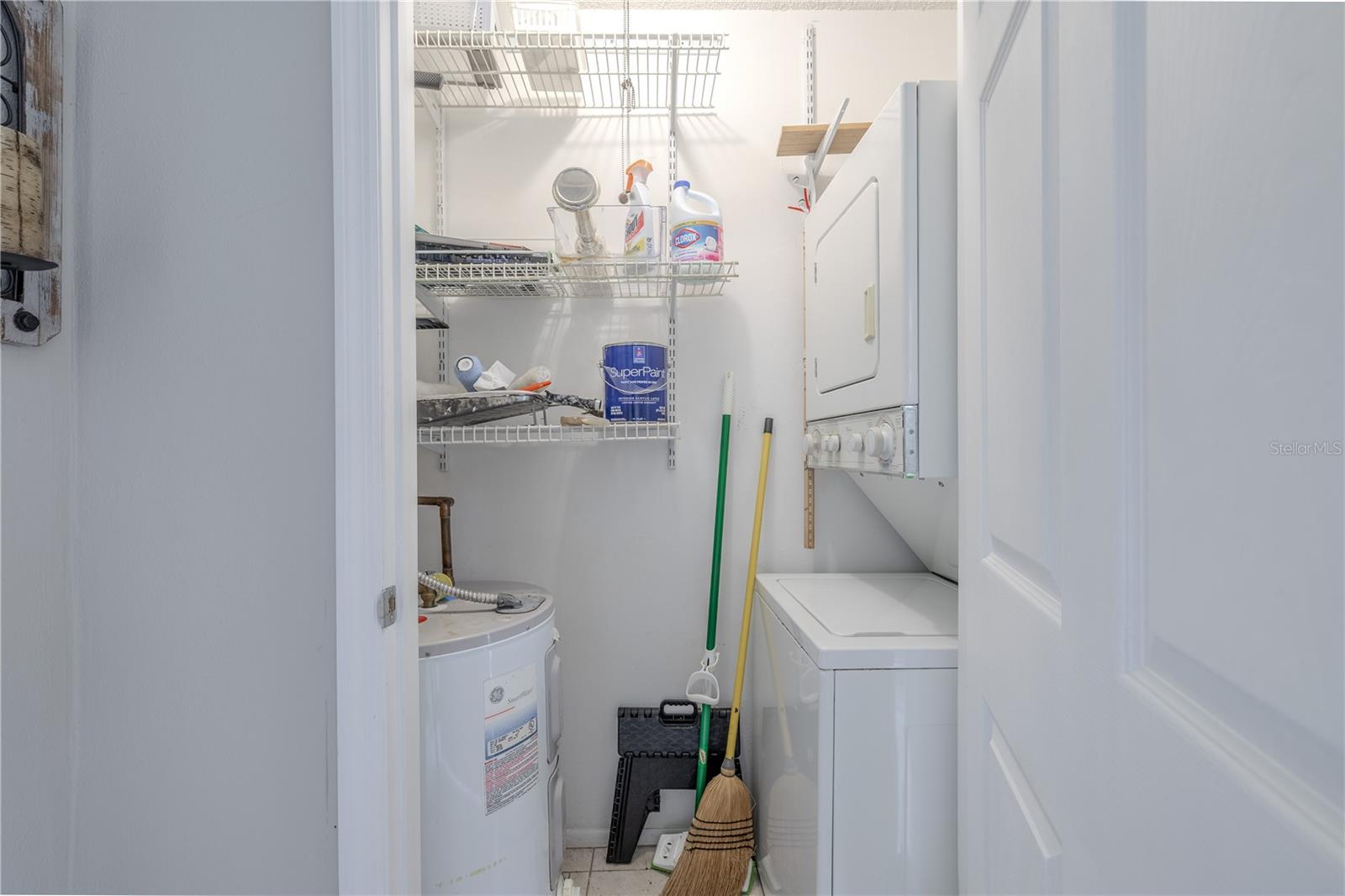Conveniently located laundry closet features a stacked washer and dryer unit, making efficient use of space while offering full functionality. Multiple wire shelves provide ample overhead storage for cleaning supplies, laundry essentials, or household tools. This compact utility area also includes access to the water heater and room for brooms, mops, and other cleaning gear—all neatly tucked behind a closet door for a clean, organized look.