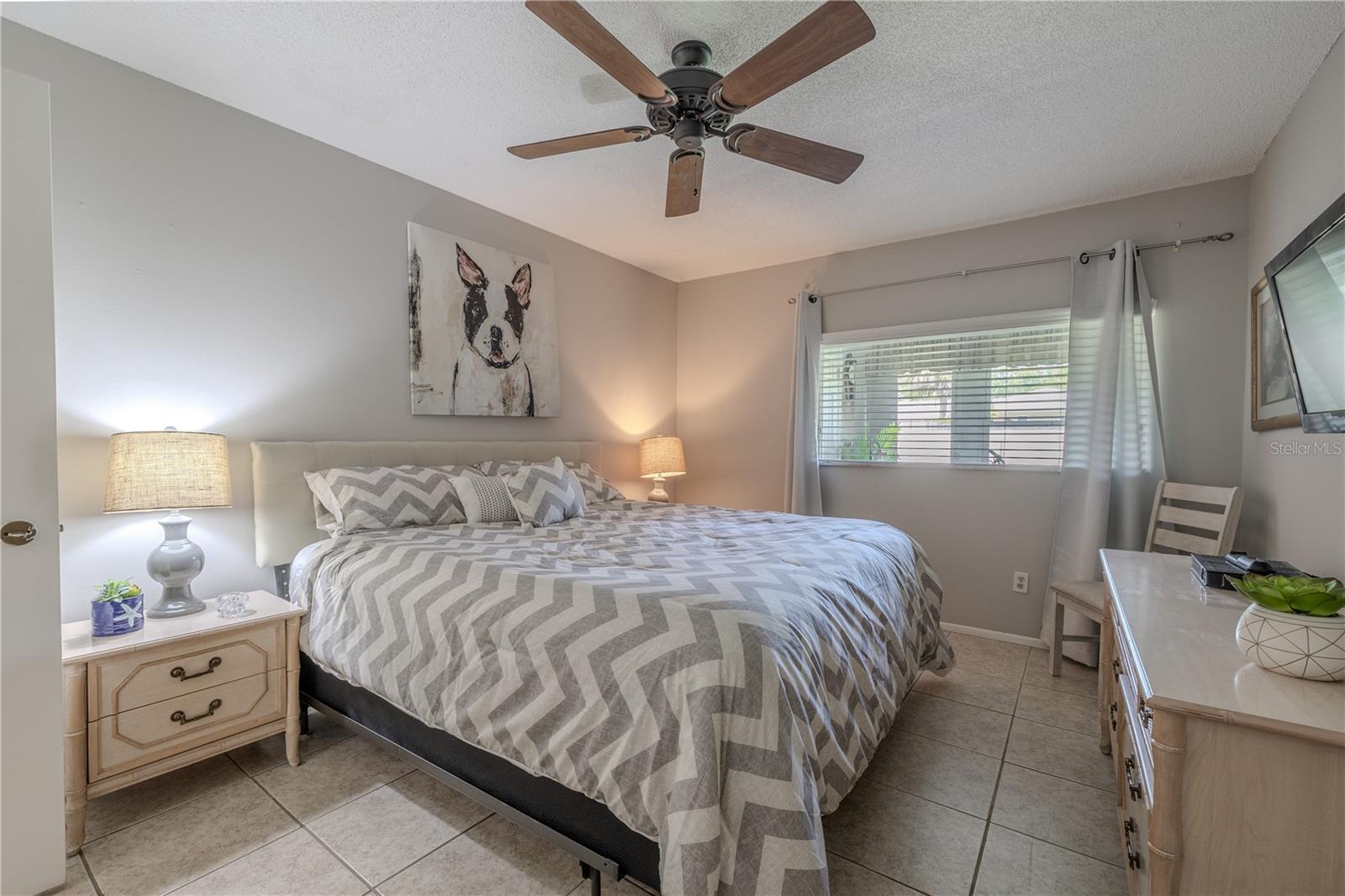 The spacious bedroom features neutral tile flooring, soft gray walls, and a large window that brings in natural light while offering privacy.