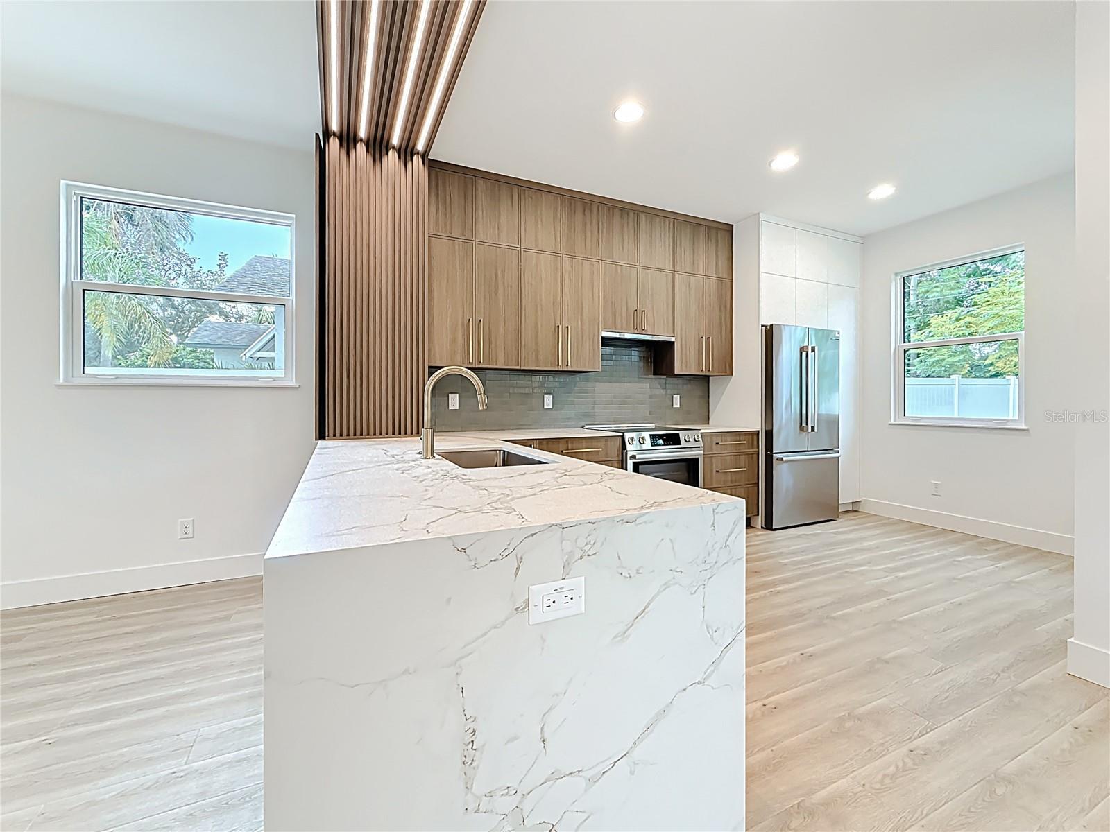 Sleek and modern kitchen featuring custom wood cabinetry, quartz waterfall countertops, stainless steel appliances, and stylish accent details — all bathed in natural light.