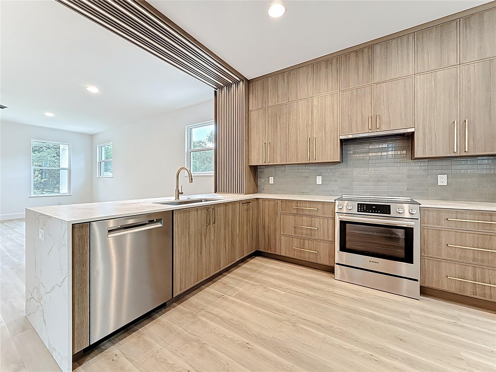 Sleek and modern kitchen featuring custom wood cabinetry, quartz waterfall countertops, stainless steel appliances, and stylish accent details — all bathed in natural light.