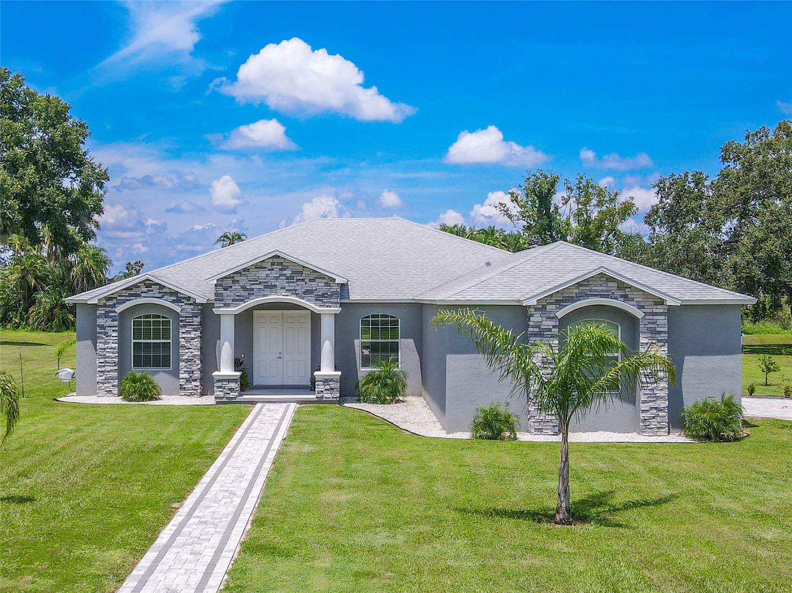 Set on a sprawling, well-manicured lot, this contemporary ranch-style home boasts a striking gray façade accented with elegant stonework and crisp white trim. A welcoming double-door entry is framed by stately columns and a covered front porch, while arched windows enhance the home’s curb appeal. The long paver walkway and matching driveway adds symmetry, leading the eye toward the home’s clean architectural lines.