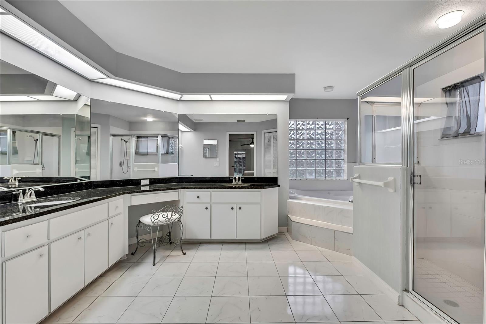 Primary bathroom has long bright double sink granite countertop vanity with vanity seating area.  Glass block wall behind the jetted garden tub provides stylish lighting and privacy.  Large glass enclosed walk in shower.  Luxurious pretty marble look tile flooring accents the white cabinets.