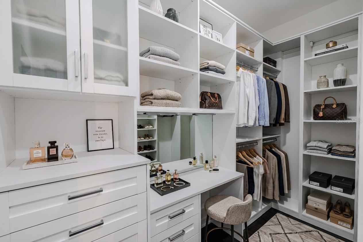 The secondary primary closet offers exceptional organization and functionality, featuring custom built-ins with open shelving, drawers, and hanging space. Frosted glass cabinet doors add a touch of elegance, while a built-in vanity with mirror and ample countertop space provides a perfect spot for getting ready. Thoughtfully designed to maximize storage and style