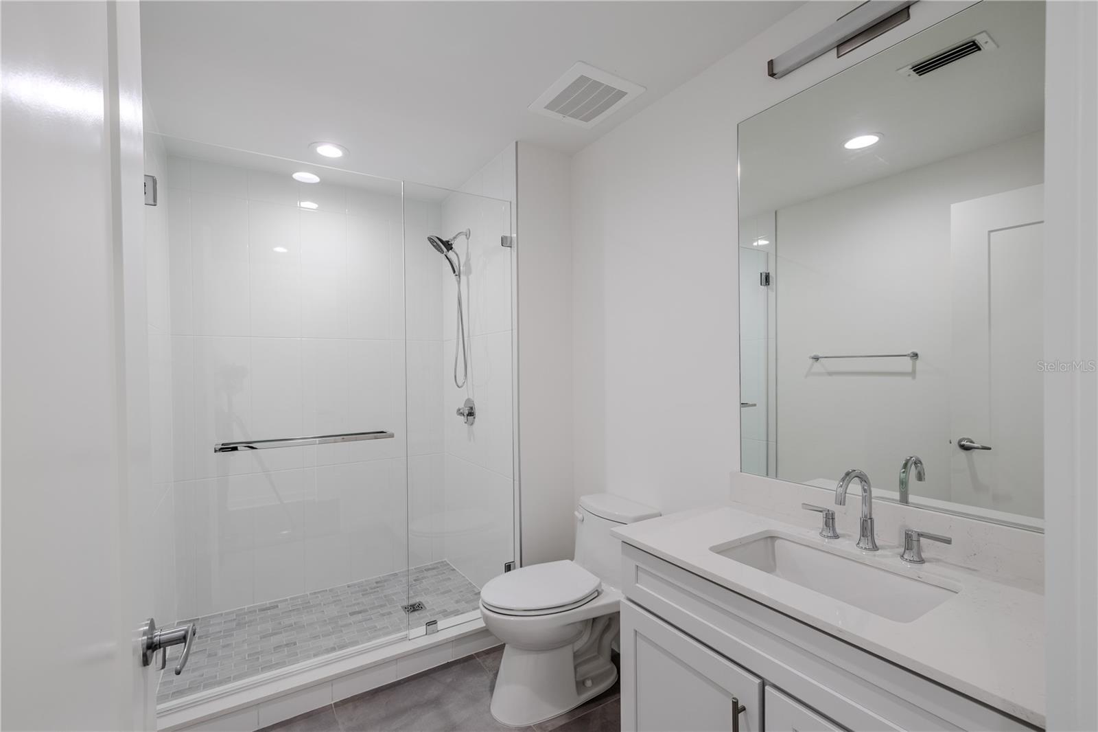 Bathroom 2 offers a sleek, contemporary design with a frameless glass-enclosed walk-in shower featuring large-format white tile and a mosaic tile shower floor. A quartz-topped vanity with under-mount sink, polished chrome fixtures, and a wide mirror with integrated lighting create a bright, spa-like atmosphere. Clean lines and neutral tones make this en-suite both elegant and functional—perfect for guests or as a private bath for the secondary bedroom.