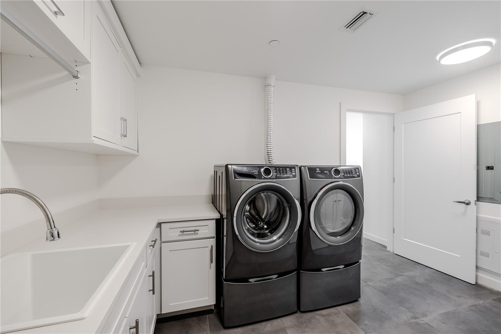 The beautifully designed laundry room features a sleek Electrolux front-load washer and dryer set, surrounded by abundant cabinetry and  countertops for folding and organization. A deep utility sink and hanging rack add everyday convenience, while modern tile flooring and recessed lighting create a clean, contemporary look. Practicality meets style in this thoughtfully planned space.