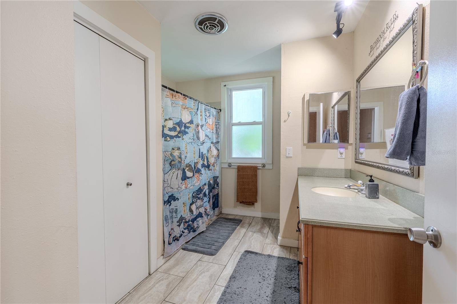 This angle of the spacious second-floor bathroom reveals a large built-in storage closet with interior shelving, ideal for towels, linens, and bathroom essentials. The room features a generous Corian countertop with single sink, a frosted window for natural light, and a wide shower area. Its ample storage and functional layout make this bathroom a highly practical part of the home.