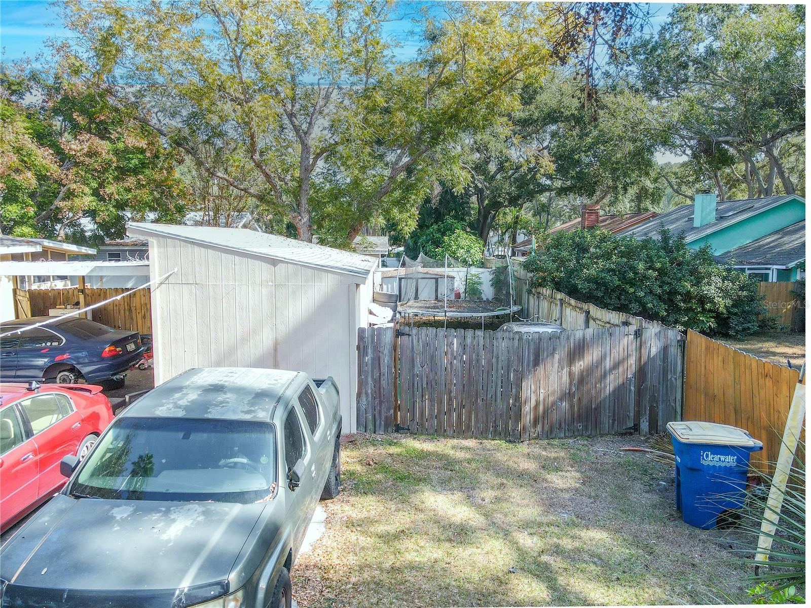 This view from the driveway showcases the rear portion of the property, including the backyard, storage shed, and fenced perimeter. The layout provides easy access to the yard and outbuildings, with plenty of space for parking additional vehicles, trailers, or equipment. Mature trees and surrounding fencing offer privacy, while the workshop adds valuable storage for tools or outdoor gear.