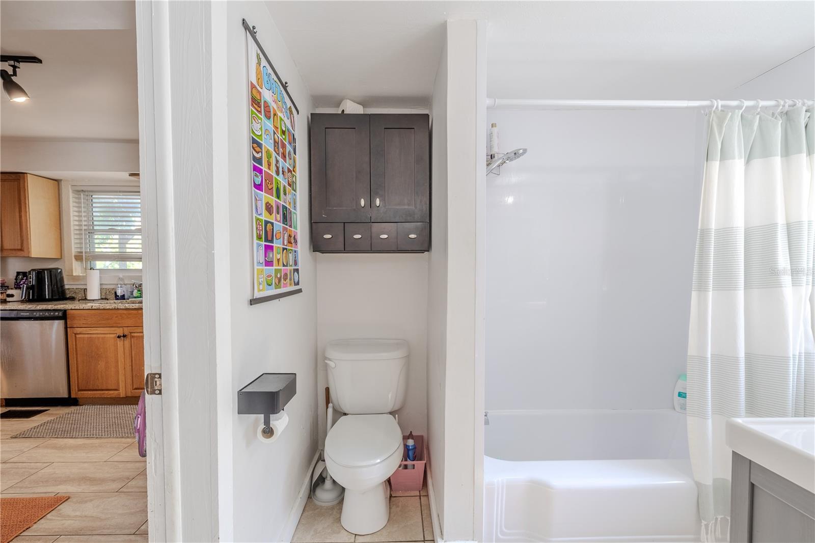 This view of the first-floor en-suite bathroom highlights its convenient secondary access point directly from the kitchen, making it easily reachable for guests and daily use. The bathroom features a shower–tub combination, a wall-mounted storage cabinet, and neutral tile flooring. Its dual-access layout—open to both the kitchen and the first-floor bedroom—adds excellent functionality to the home’s main level.