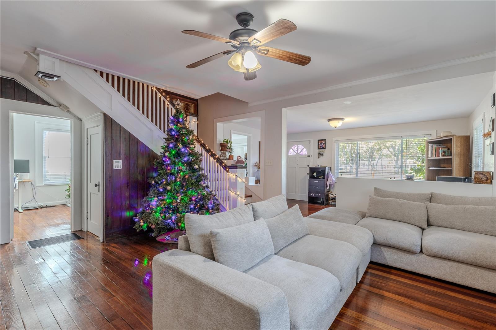 This angle of the living room highlights the home’s functional layout, featuring convenient access to the first-floor bedroom through the doorway on the left. The charming under-stair door provides additional storage space, maximizing usability while adding character to the home’s historic design. Original wood floors shine throughout, complemented by warm natural light and an overhead ceiling fan for added comfort. This spacious living area offers a welcoming flow that connects easily to adjacent rooms and everyday living spaces.