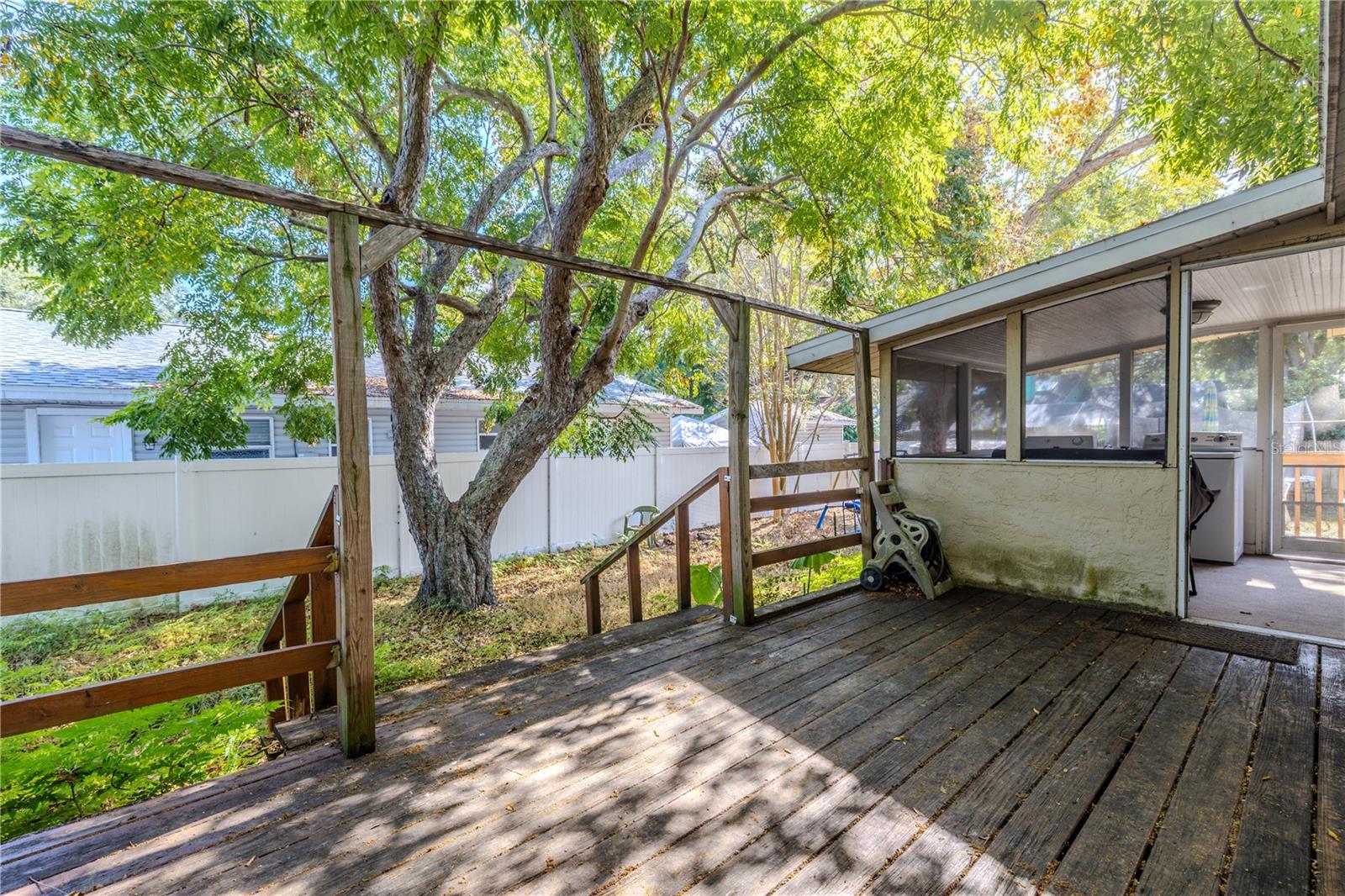 This view from the storage area shows the spacious wooden deck extending toward the screened-in porch, offering a seamless transition between indoor and outdoor living. Mature shade trees create a peaceful, natural backdrop, and the fully fenced backyard adds privacy and security. This versatile outdoor area is ideal for relaxing, entertaining, or enjoying weekend projects, with the convenience of the attached storage room just steps away.