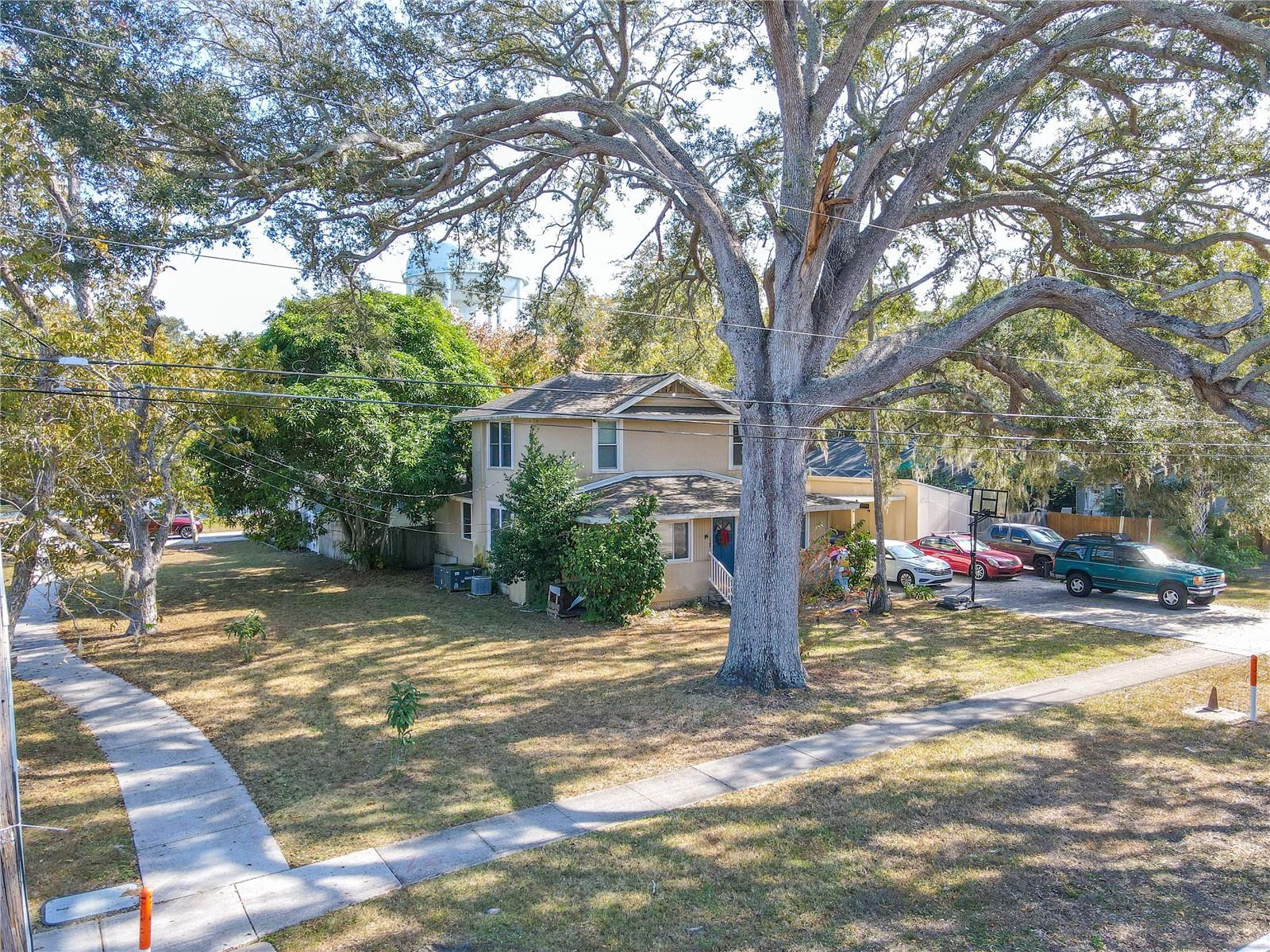 This corner lot view beautifully showcases the home’s prominent placement and expansive yard space, framed by stunning mature shade trees that add charm and character. Sidewalks border both street fronts, enhancing walkability and curb appeal. The large driveway and carport sit to the right, while the home itself is set back comfortably on the lot, offering privacy and a welcoming presence. A standout perspective that highlights the property’s generous outdoor space and desirable corner location.