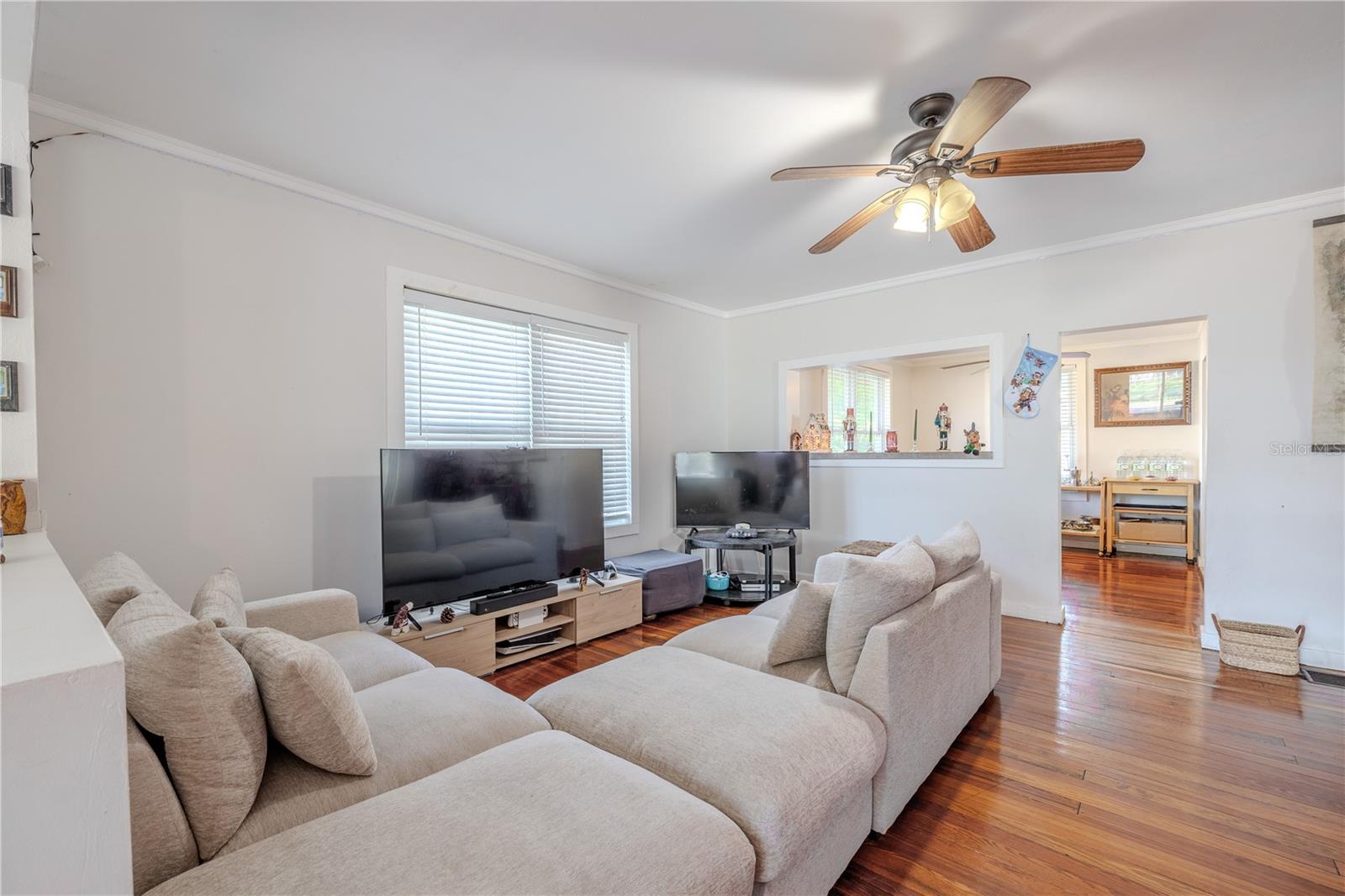 This deeper view into the living room highlights the home’s warm and inviting atmosphere, with beautifully finished wood floors and generous natural light filling the space. A central ceiling fan enhances comfort, while the open layout provides an easy flow into the dining room just beyond the passthrough and doorway. The room offers plenty of space for multiple seating arrangements, making it ideal for relaxing, gathering, or entertaining. The glimpse of the staircase adds a touch of character, reflecting the timeless charm of this historic home.