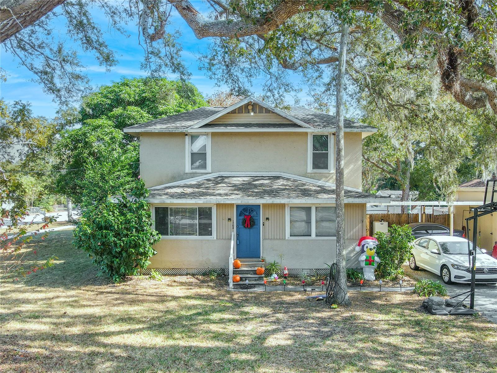 This charming historic 1918 home stands proudly on a spacious corner lot, shaded by beautiful mature oaks that frame its classic two-story profile. The inviting façade features a bold blue front door and large picture windows that brighten the interior with natural light. The expansive yard and sidewalks surrounding the property make it ideal for easy neighborhood strolls and outdoor enjoyment. A long, wide driveway provides ample parking, leading to a covered carport with an attached workshop—perfect for storage, hobbies, or weekend projects. Thoughtful seasonal touches and established landscaping enhance the warm, welcoming curb appeal of this unique historic gem.