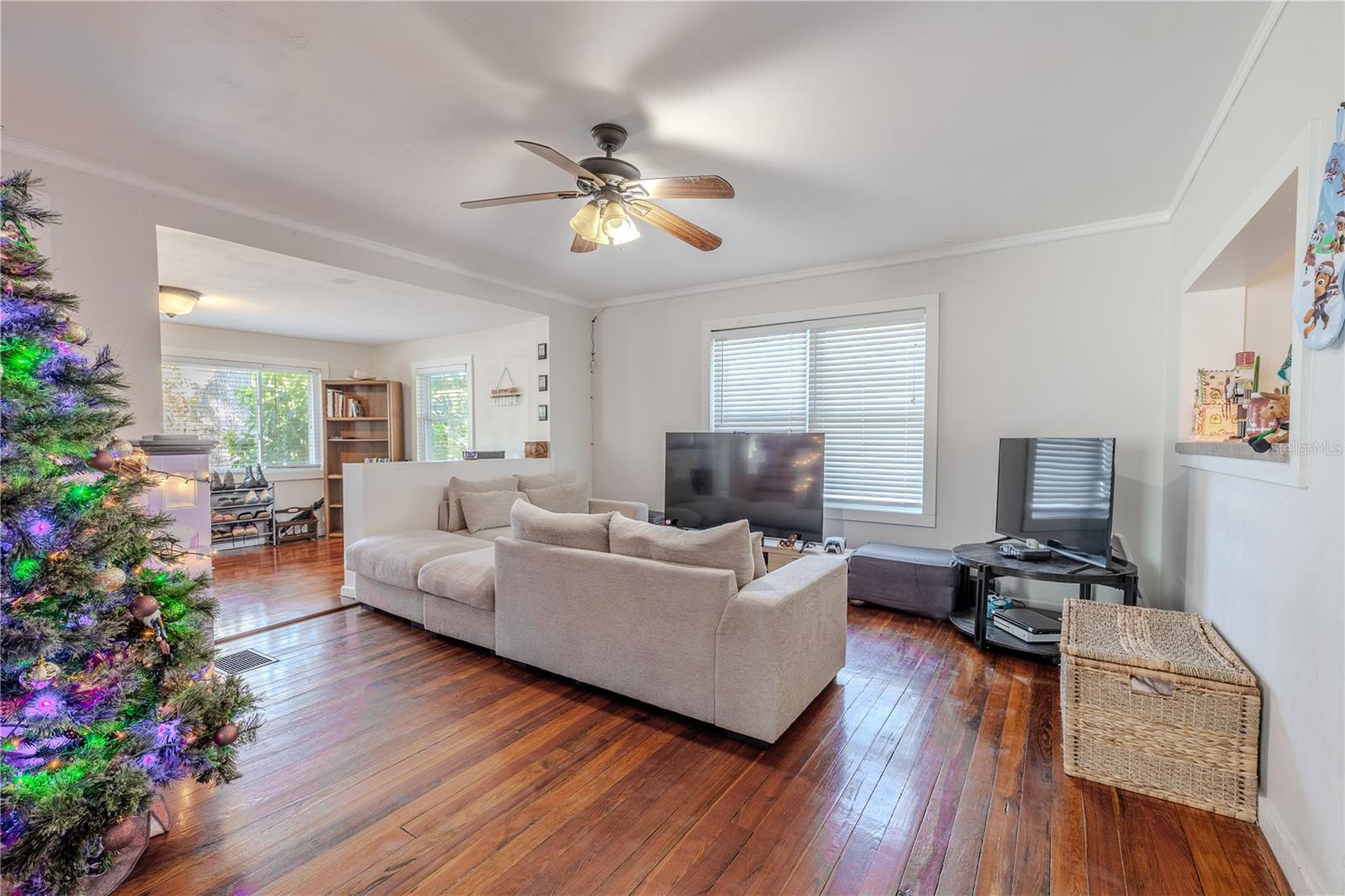 This full view of the living room highlights an expansive, light-filled space featuring beautifully preserved original wood floors and generous room for multiple seating arrangements. Large windows invite natural light throughout the day, enhancing the warm and inviting atmosphere. A central ceiling fan provides added comfort, and the open layout flows seamlessly into the adjacent flex space and dining area, creating a connected main living level ideal for entertaining or everyday family living. The room’s size, charm, and versatility make it a standout feature of this historic home.