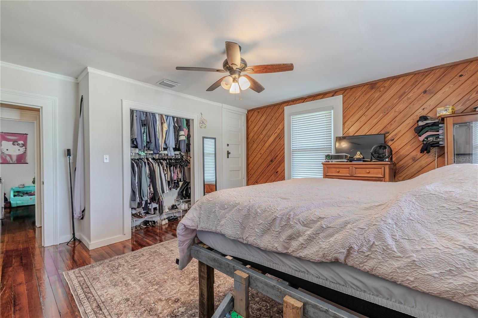 This view of the second-floor master bedroom highlights the spacious built-in closet, offering generous hanging space and easy organization. Additional shelving and storage to the right provide even more room for clothing or personal items. The room features warm wood accent paneling, standard crown molding, and beautifully maintained wood floors, all enhanced by natural light from multiple windows. This comfortable and well-appointed master bedroom offers both charm and practicality.