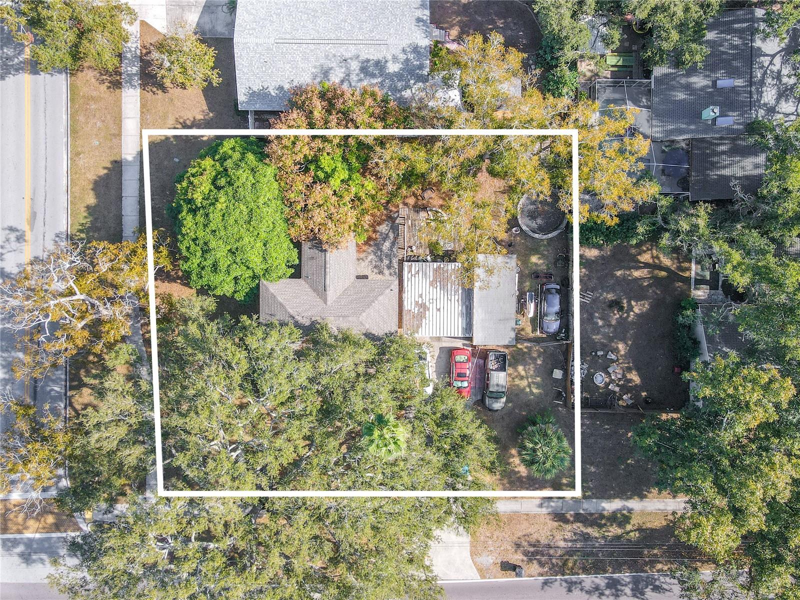 This expansive aerial view highlights the generous corner lot that sets this 1918 historic home apart. The outlined property showcases the full scope of the land, including mature shade trees, wide side and front yards, and ample open space surrounding the residence. The layout features a long driveway leading to the carport and workshop area, with additional parking options clearly visible. Nestled among lush greenery, the home enjoys excellent privacy while still being conveniently bordered by sidewalks on multiple sides—ideal for neighborhood strolls. This overhead perspective beautifully captures the property’s size, usability, and overall potential.