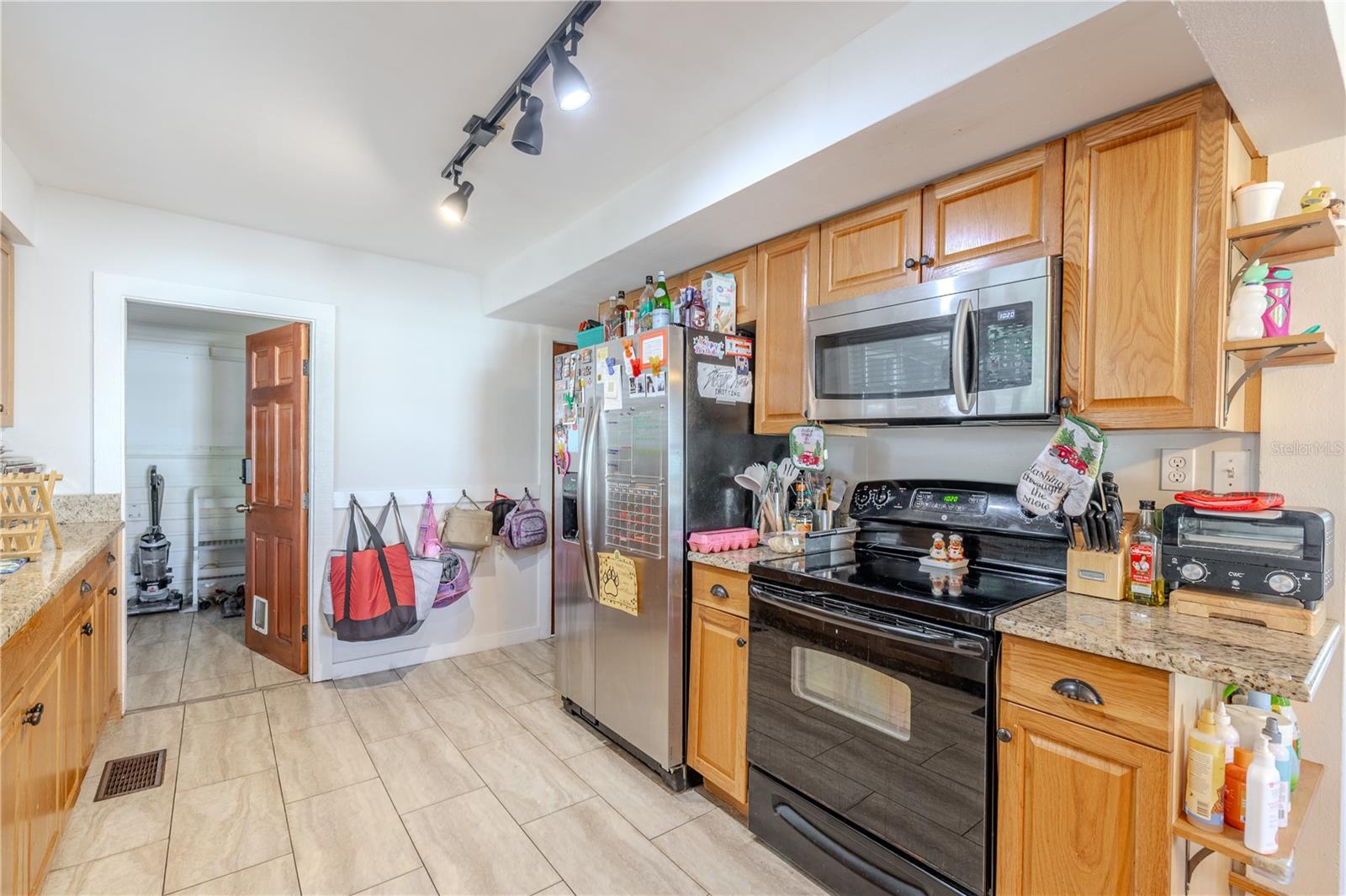 This additional kitchen view highlights the updated appliance package, including a stainless-steel refrigerator, built-in microwave, and black electric range. Warm wood cabinetry and granite countertops provide ample storage and workspace, while overhead track lighting ensures bright, even illumination. The tile flooring offers durability and easy upkeep, and the open doorway leads directly into the spacious walk-in pantry for added convenience. This well-appointed kitchen combines functionality with everyday comfort.